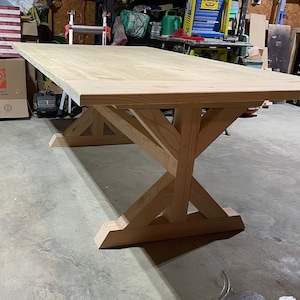 Puede incluir: Una mesa de madera grande y sin terminar con una tapa rectangular y una base en forma de X única. La mesa está hecha de madera de color claro y tiene un aspecto natural y rústico. La mesa está sobre un suelo de hormigón.