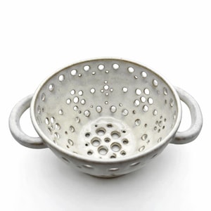 May include: A white ceramic colander with numerous small holes and two loop handles. The bowl has a rounded shape and is designed for draining liquids from food items. The colander is on a white background.
