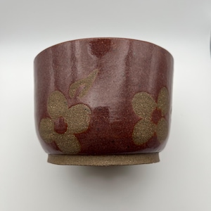 May include: A handmade ceramic bowl with a reddish-brown glaze and a textured, unglazed base. The bowl features a floral design in a lighter, golden-brown color. The interior is a darker shade of the same reddish-brown.