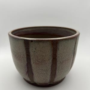 May include: A handmade ceramic bowl with a textured, speckled finish. The bowl is a muted green color with vertical brown stripes and a dark red rim. The bowl has a rounded shape and is suitable for serving food.