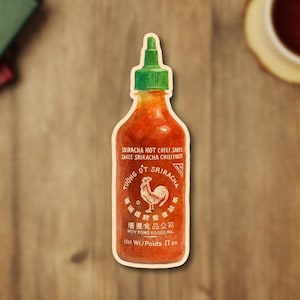 May include: A sticker of a Sriracha hot chili sauce bottle with a green cap. The label reads "SRIRACHA HOT CHILI SAUCE" and "HUY FONG FOODS INC." The bottle contains 17 oz of sauce.