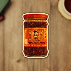 May include: A sticker of a jar of Lao Gan Ma Spicy Chili Crisp. The jar has a red lid and a label with Chinese characters and the text "SPICY CHILI CRISP". The net weight is 7.41 oz (210g).