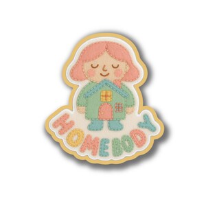 May include: A felt patch featuring a person-shaped design with a house-shaped body. The person has pink hair and is wearing a green house. The word "HOMEBODY" is stitched in colorful letters below the figure. The patch has a yellow border.