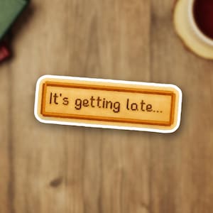 May include: A rectangular sticker with the text "It's getting late..." in a pixelated font. The text is inside a brown rectangle with a white border. The sticker is on a wooden surface.