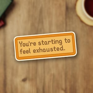 May include: A rectangular sticker with the text "You're starting to feel exhausted." in a pixelated font. The sticker has a light orange background with a darker orange border and a white outline. The sticker is on a wooden surface.
