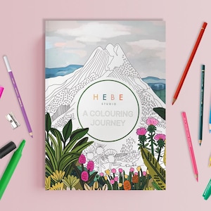 May include: A coloring book titled "A Colouring Journey" with a mountain landscape illustration, surrounded by colored pencils, a highlighter, and a sharpener. The book cover features the text "HEBE Studio".
