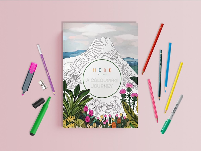 May include: A coloring book titled "A Colouring Journey" with a mountain landscape illustration, surrounded by colored pencils, a highlighter, and a sharpener. The book cover features the text "HEBE Studio".