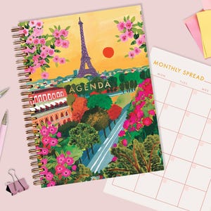 Peut inclure: Un agenda à spirales avec une illustration colorée de la Tour Eiffel, de fleurs et d'un coucher de soleil. Le mot "AGENDA" est imprimé sur la couverture. Un calendrier mensuel et des stylos roses sont également visibles.