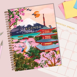 Peut inclure: Agenda à spirales avec un motif de paysage japonais. La couverture présente une pagode, le mont Fuji et des fleurs de cerisier. Le mot "AGENDA" est imprimé sur la couverture. Des stylos roses, une pince à reliure et des post-it sont sur la surface rose.