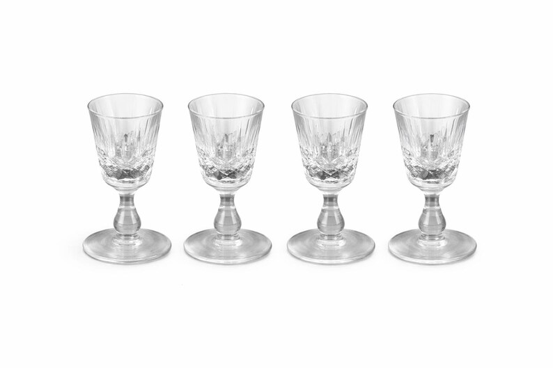 May include: Four clear crystal shot glasses with a cut-glass design. Each glass has a round base, a stem, and a flared rim. The glasses are arranged in a row against a white background. Ideal for serving spirits.