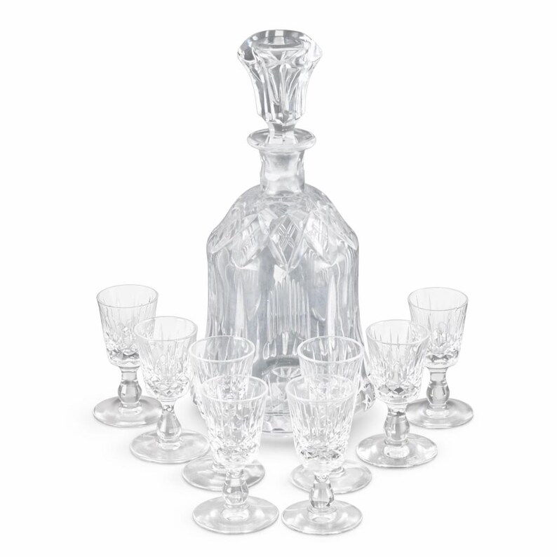 May include: A clear crystal drinks decanter with a stopper, accompanied by eight matching shot glasses. The decanter and glasses have a cut glass pattern. The set is displayed against a white backdrop.