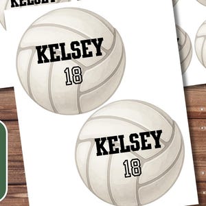 May include: Sheets of volleyball-themed stickers with the name "Kelsey" and the number "18" printed on them. The stickers are white with a volleyball design and black text. The image also includes a coffee cup, pen, and greenery.