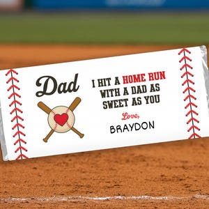May include: A white candy bar wrapper with baseball-themed graphics and text. The design includes a baseball with a heart, crossed bats, and red stitching. Text reads "Dad, I hit a home run with a dad as sweet as you, Love, Braydon."