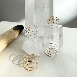 May include: A collection of spiral rings in gold and silver tones. The rings are displayed on clear quartz crystal formations, with a large crystal point in the background. A bundle of Palo Santo wood is on the left.