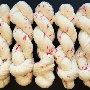 Op de afbeelding: Vijf strengen crèmekleurig garen met levendige rode en roze spikkels. Het garen is strak getwist, waardoor een textuur ontstaat. De strengen zijn in een rij gerangschikt tegen een donkere achtergrond, waardoor de kleur en details van het garen worden getoond.