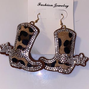May include: A pair of dangle earrings shaped like cowboy boots with a brown and black leopard print pattern. The boots are outlined in clear rhinestones and feature a rhinestone-encrusted spur. The earrings are attached to a white card labeled Fashion Jewelry.