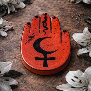 May include: A red, hand-shaped decorative item with black symbols, including a crescent moon and a cross. The tips of the fingers have black dots. White lilies with dark spots surround the item on a wooden surface.