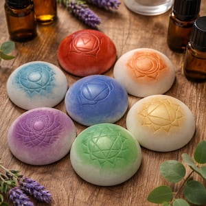 May include: Seven colorful chakra stones with embossed symbols are arranged on a wooden surface. The stones are in shades of red, orange, yellow, green, blue, indigo, and purple. Essential oil bottles, lavender sprigs, and a lit candle are also present.