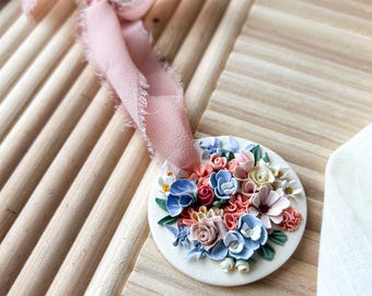 May include: A white ceramic ornament featuring a colourful floral design. The ornament includes a variety of flowers in shades of pink, blue and yellow. The ornament is attached to a pink ribbon.