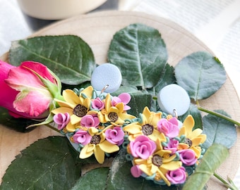 May include: A pair of dangle earrings featuring a light blue circular stud attached to a cluster of sculpted yellow sunflowers and pink roses. The floral arrangement is detailed with small brown centres and green accents, creating a textured, multi-layered appearance on each earring.