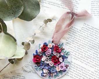 May include: A round, white ornament featuring a cluster of red, pink, and purple polymer clay flowers. The flowers are arranged in a bouquet shape and have a realistic appearance.