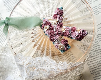 May include: A delicate, handmade letter "K" ornament crafted from pink and white fabric roses. The ornament is displayed on a clear glass plate with a green ribbon.