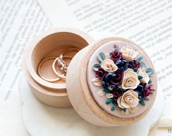 May include: A round wooden ring box with an open lid. The top of the lid features a cluster of sculpted cream, burgundy, and dark blue flowers with green leaves. Inside the box, two thin gold-toned metal rings are visible resting on the base.
