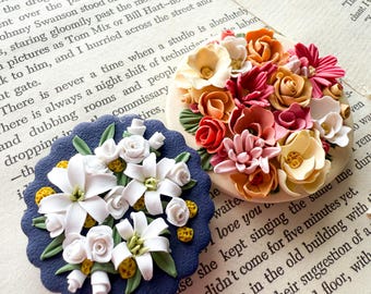 May include: Two decorative floral arrangements. One is a round, cream base with a bouquet of orange, red, and pink flowers. The other is a navy blue, scalloped-edge base with white flowers and yellow accents. Both are on a book page.