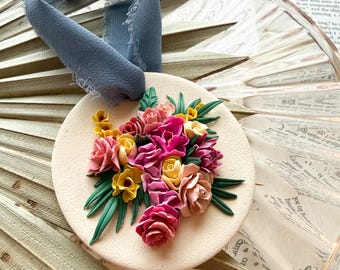 May include: A circular cream-coloured ornament featuring a sculpted floral arrangement in shades of pink, yellow, and magenta with green leaves. A frayed dusty blue ribbon is tied through a hole at the top for hanging the decorative piece.
