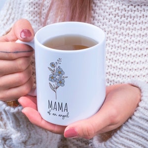 May include: White ceramic mug with a floral design and the text "MAMA of an angel". The mug is filled with a light brown liquid.
