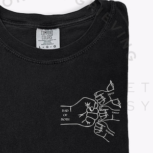May include: Black t-shirt with a white graphic of clasped hands and the words "DAD OF BOTH". The shirt also features the text "LOVE", "JOY", and "BLESSINGS". The shirt is from the brand "COMFORT COLORS".