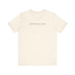 May include: Cream-colored t-shirt with the text "remembering you, always" and "PREGNANCY & INFANT LOSS AWARENESS" printed in a delicate, light gray font. The shirt is made of soft material.