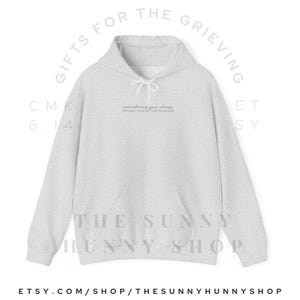 May include: A light gray hoodie with a drawstring hood and a front pocket. The text "remembering you, always" is printed on the chest. The words "GIFTS FOR THE GRIEVING" and "THE SUNNY HUNNY SHOP" are also visible.