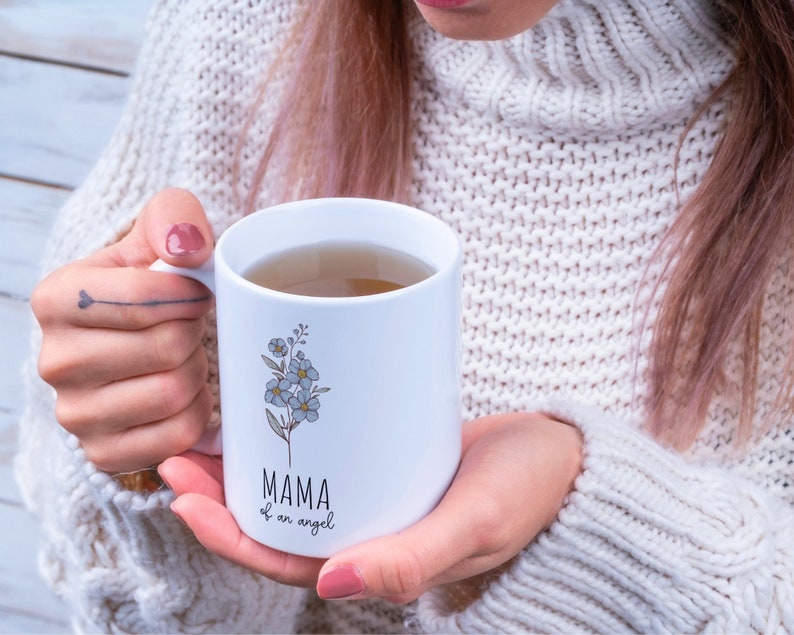May include: White ceramic mug with a floral design and the text "MAMA of an angel". The mug is filled with a light brown liquid.