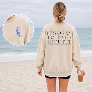 May include: A beige crewneck sweatshirt featuring a pink awareness ribbon intertwined with blue flowers on the front left chest. The back displays the text IT'S OKAY TO TALK ABOUT IT and pregnancy & infant loss awareness with a small ribbon graphic.