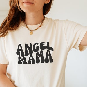 May include: A white t-shirt with the text "Angel Mama" in black, stylized lettering. The shirt has a relaxed fit and short sleeves.