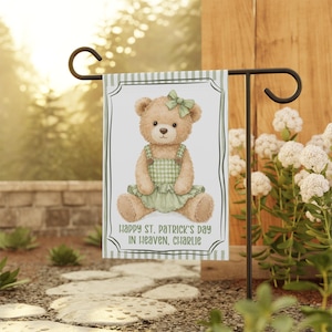 May include: A garden flag featuring a watercolor illustration of a teddy bear wearing a green gingham dress and bow. The flag has the text "Happy St. Patrick's Day in Heaven, Charlie" and is displayed on a black metal stand.