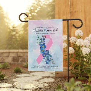 May include: A garden flag with a watercolor design. It features a pink ribbon, blue flowers, and text that reads "During October, we remember Charlotte Marion Krol and all the babies taken too soon." The flag is for Pregnancy & Infant Loss Awareness.