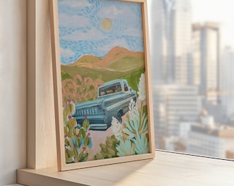 May include: A framed art print featuring a light blue Chevrolet pickup truck driving through a desert landscape. The artwork includes cacti, mountains, and a bright sun in a blue sky. The frame is made of light-colored wood.