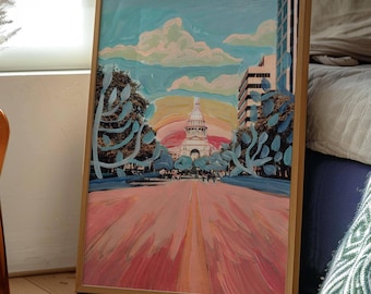 May include: A framed art print featuring a painterly depiction of the Texas State Capitol building. The scene shows a pink-toned street leading toward the white domed structure under a blue sky with clouds, flanked by trees and city buildings.