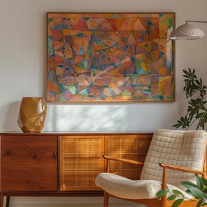 May include: A rectangular framed abstract painting with geometric shapes in shades of orange, blue, and pink hangs above a wooden sideboard. To the left sits a faceted mustard-colored vase, and to the right is a textured cream-colored armchair.