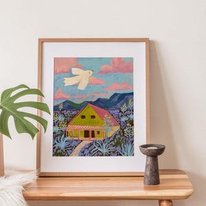 May include: A framed art print of a landscape painting. The artwork shows a yellow house with a pink roof, a white bird in flight, and a path leading to the house. The sky is blue with pink clouds, and the scene is surrounded by purple and green foliage.