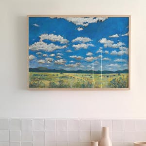 May include: A rectangular landscape painting in a light wood frame depicting a vast field with yellow and green grasses under a bright blue sky filled with numerous white, fluffy clouds. Dark hills are visible along the distant horizon line.