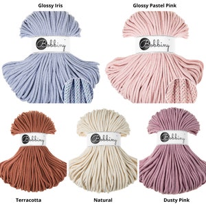 May include: A collection of colorful Bobbiny macrame cords in various shades. The colors include Glossy Iris, Glossy Pastel Pink, Terracotta, Natural, and Dusty Pink. Each spool is labeled with the Bobbiny brand name.