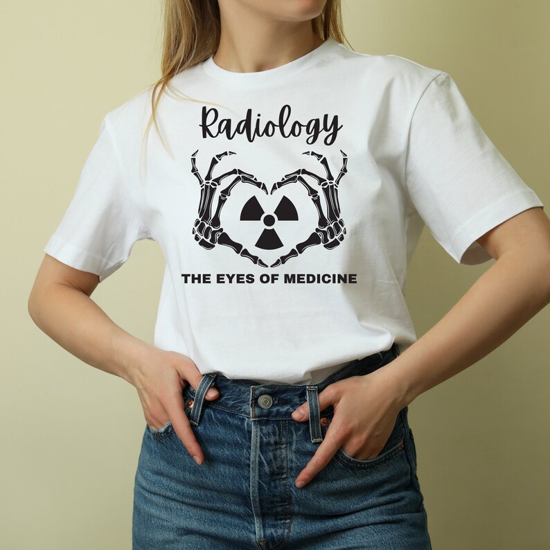 May include: White t-shirt with black text that reads "Radiology The Eyes of Medicine". The text is surrounded by a heart shape made of two skeleton hands.