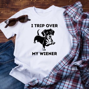 Op de afbeelding: Wit T-shirt met de tekst "I TRIP OVER MY WIENER" en een zwarte teckelafbeelding. De afbeelding bevat een zonnebril, denim en een geruit overhemd, alles op een houten achtergrond.