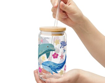 May include: A clear glass reusable water bottle with a bamboo lid and a straw. The bottle has a colorful design featuring dolphins, whales, and flowers. The text "Aloha" is printed on the bottle.
