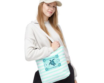 May include: A white tote bag with horizontal light blue stripes. The bag features a central square design with a blue sea turtle and floral accents. The bag is carried over the shoulder.