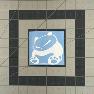 May include: White vinyl sticker of a cartoon animal with a grumpy expression. The animal is sitting with its paws crossed and has a blue background.