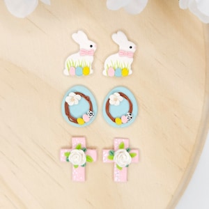 May include: Four pairs of Easter-themed clay earrings. Two pairs feature white bunnies with pink bows and pastel colored eggs. One pair features blue wreaths with white flowers and pastel colored eggs. One pair features pink crosses with white flowers and green leaves.